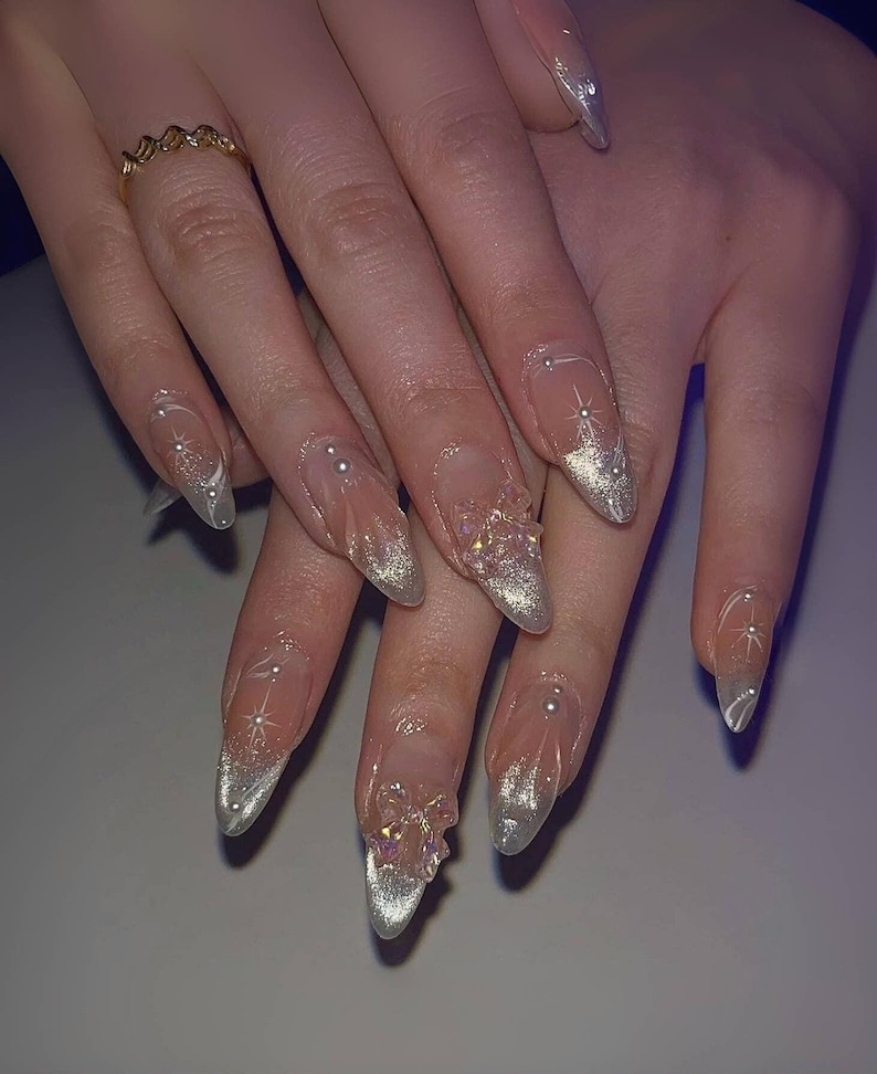 May include: Close-up of a hand with long, almond-shaped nails. The nails are painted with a clear, iridescent polish and decorated with silver glitter, rhinestones, and white designs.