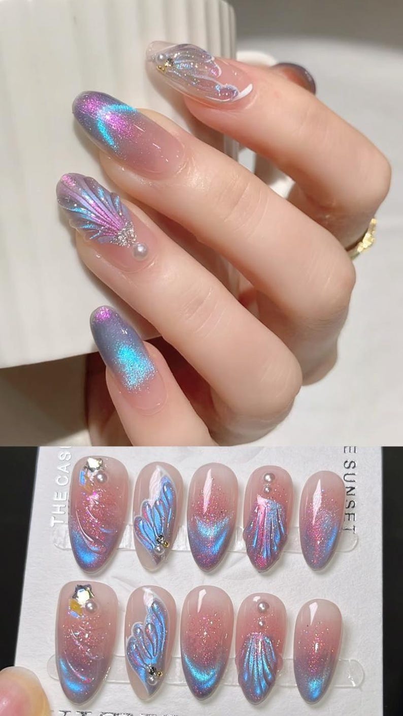 K&ouml;nnte beinhalten: K&uuml;nstliche N&auml;gel mit einem Farbverlauf in Rosa, Lila und Blau. Einige N&auml;gel weisen ein Muscheldesign mit schillerndem Glitzer und kleinen Perlenakzenten auf. Die N&auml;gel werden auf einer Hand und in einem Set gezeigt.