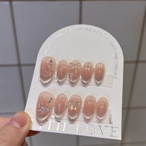 May include: A set of 10 false nails with a clear base and glitter accents. Some nails have a small bow design. The nails are displayed on a white card with the text "A casual encounter is more gentle than the sunset!" and "With Love".