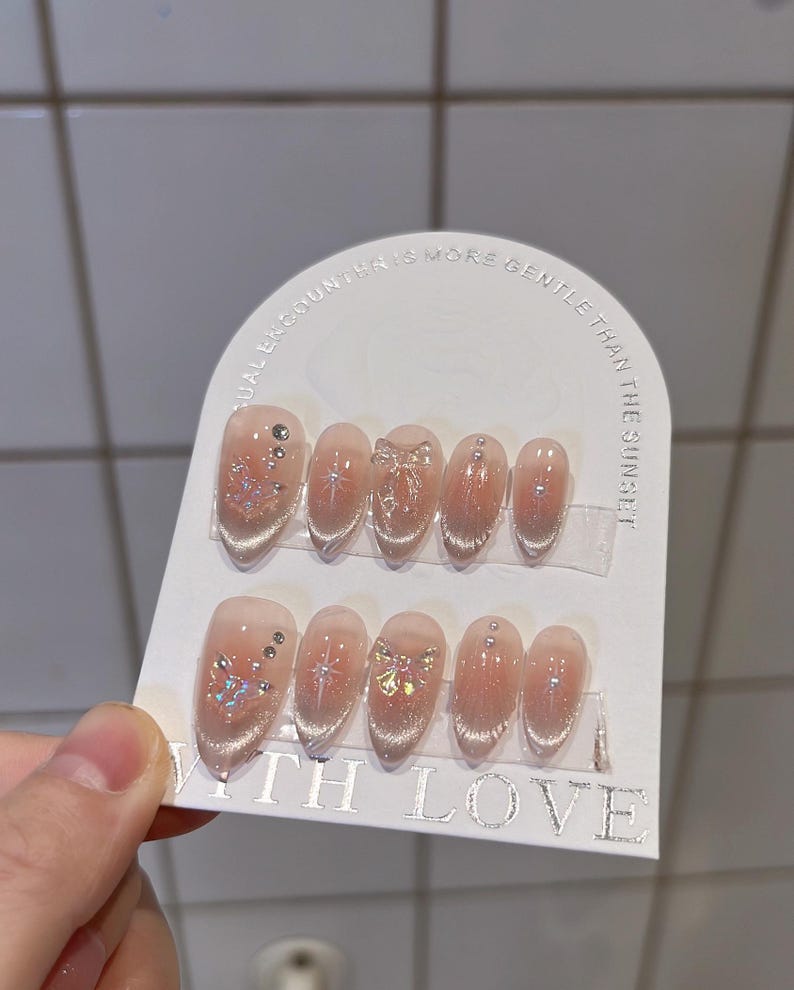 May include: A set of 10 false nails with a clear base and glitter accents. Some nails have a small bow design. The nails are displayed on a white card with the text "A casual encounter is more gentle than the sunset!" and "With Love".
