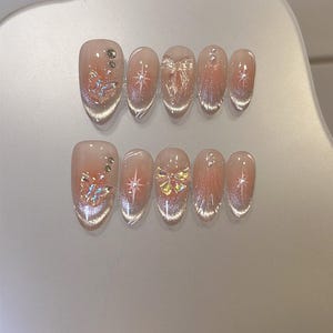 May include: Set of ten clear press-on nails with different designs. Each nail features a sparkly finish and a small, decorative accent. The designs include a butterfly, a star, and a bow.