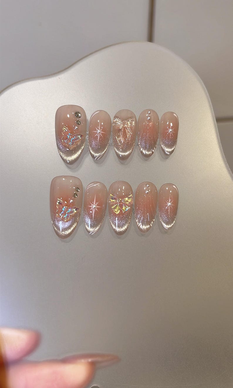 May include: Set of ten clear press-on nails with different designs. Each nail features a sparkly finish and a small, decorative accent. The designs include a butterfly, a star, and a bow.