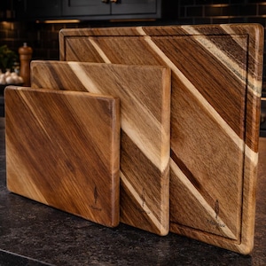 May include: Three wooden chopping boards of different sizes, displayed together. They are made from light and dark brown wood with a diagonal grain. The largest board has a juice groove. Each board has a logo.