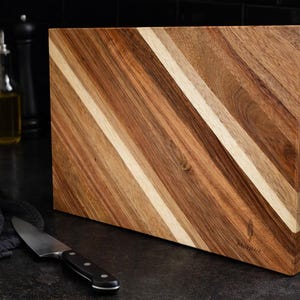 May include: A rectangular wooden chopping board with a diagonal striped pattern in shades of brown and tan. A stainless steel knife with a black handle rests on the dark surface. Kitchenware.