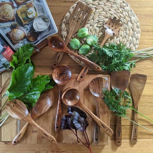 Może przedstawiać: Widok z góry na różne drewniane przybory kuchenne, w tym łyżki i szpatułki, ułożone na drewnianej desce do krojenia i plecionym podkładce. Obecne są również świeże warzywa, takie jak brukselka i marchew.