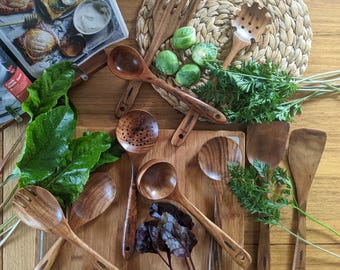 Puede incluir: Vista cenital de varios utensilios de cocina de madera, incluyendo cucharas y espátulas, dispuestos sobre una tabla de cortar de madera y un salvamanteles tejido. También hay verduras frescas, como coles de Bruselas y zanahorias.