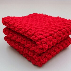 May include: A stack of three red crocheted dishcloths. The dishcloths are made with a simple stitch pattern and are perfect for cleaning up spills and messes in the kitchen.