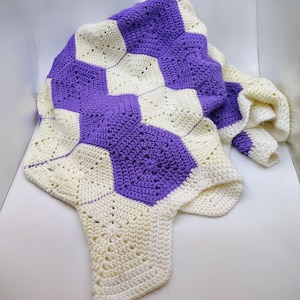 May include: A crocheted blanket with a geometric pattern of purple and cream-coloured hexagons. The blanket is folded, displaying the texture and design of the yarn. The overall aesthetic is cosy and handcrafted.