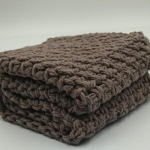 Puede incluir: Un conjunto de cuatro paños de cocina de color marrón hechos a crochet. Los paños están hechos con un punto texturizado y están doblados cuidadosamente.