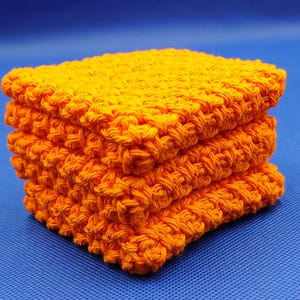 May include: A stack of four bright orange, hand-crocheted square washcloths. The washcloths are made of thick yarn, with a textured, raised pattern. The background is a solid blue.