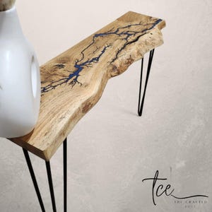 May include: A wooden console table with a live edge and natural wood grain. It has a blue resin lightning bolt design. The table has black hairpin legs. A white vase is partially visible on the left. The table's length is approximately 100 cm.