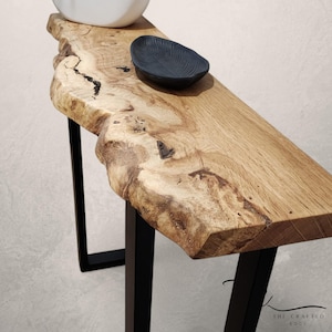 Puede incluir: Una consola de borde vivo con acabado de madera natural. La mesa presenta una parte superior gruesa e irregular sostenida por patas de metal negro. Un pequeño cuenco azul oscuro descansa sobre la superficie. La mesa mide unos 122 cm de largo.