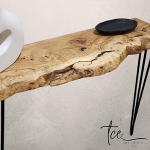 Könnte beinhalten: Eine Konsole aus Holz mit lebendiger Kante und schwarzen Haarnadelbeinen. Der Tisch hat eine natürliche Holzmaserung und eine dunkle, ovale Schale. Auf der linken Seite des Tisches befindet sich eine weiße Vase. Der Tisch ist ca. 122 cm lang.