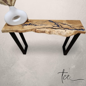 Puede incluir: Banco de madera de borde vivo con incrustaciones de resina azul, sostenido por patas de metal negro. La madera tiene un color marrón claro natural con un patrón de vetas único. Un jarrón blanco con una abertura circular está sobre el banco. The Crafted Edge.
