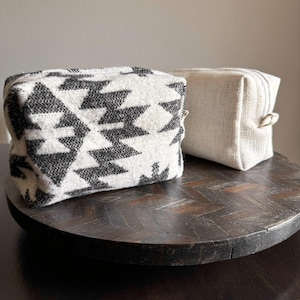 May include: Two small, rectangular fabric pouches. The pouch on the left has a black and white geometric pattern. The pouch on the right is a solid beige color. Both pouches are sitting on a dark wooden surface.