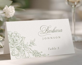 May include: A white folded place card with a textured surface featuring a sage green floral illustration on the left side. The text reads Barbara JOHNSON and Table 5 in a dark grey serif font. The card is positioned upright on a flat surface.