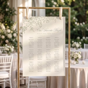 May include: A cream-coloured seating chart sign suspended from a gold metal frame by two white ribbons. The sign features a floral line drawing at the top, the text "Our Favorite People" and "Please Find Your Seat," and a list of twelve tables.