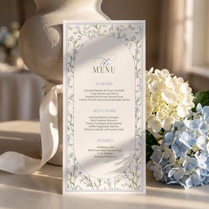 Może przedstawiać: Prostokątna karta menu z centralną owalną ramką i niebieskimi ilustracjami kwiatowymi. Tekst zawiera nagłówki The Menu, Starters, Main Course i Desserts oraz listę potraw, takich jak wędzony łosoś, koktajl z krewetek i tort weselny.