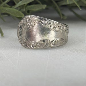Può includere: Un anello color argento con un design vintage ornato. L'anello presenta un motivo inciso dettagliato con una forma ovale centrale e abbellimenti a spirale. L'anello è probabilmente realizzato in argento sterling o metallo simile.