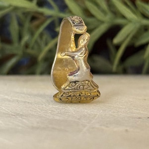 Puede incluir: Un anillo de metal dorado con un diseño decorativo. El anillo presenta una figura esculpida de una persona con un vestido fluido, con detalles intrincados. El anillo tiene una estética vintage o antigua.