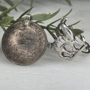 May include: A silver pendant made from a coin with the text "Vente Cente" and a ring with a detailed bird design. The coin pendant has a loop for a chain. The ring features a bird perched on a branch.