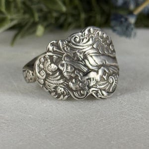May include: Ornate silver ring featuring detailed floral and scrollwork designs. The ring has a wide, decorative face with a raised central motif and a textured band. The ring's design is intricate and vintage-inspired.