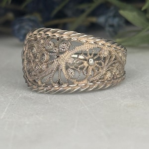 Puede incluir: Un anillo de filigrana plateado con intrincados diseños florales y de volutas. El anillo presenta un patrón calado detallado con una flor central y pequeños acentos redondos. La banda tiene una textura trenzada.