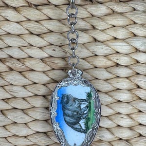 May include: Silver-toned keychain with a decorative oval pendant. The pendant features a hand-painted image of a gray koala bear on a blue and green background, framed by an ornate silver border. The keychain is attached to a woven background.
