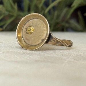 May include: Antique gold-toned ring with a circular setting and a twisted band. The setting features a small, textured gold detail in the center. The ring is set against a neutral background with blurred greenery.
