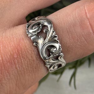 Könnte beinhalten: Ein silberfarbener Ring mit einem kunstvollen, wirbelnden Filigranmuster. Der Ring weist detaillierte Schnörkel und florale Motive auf, wodurch eine Vintage- oder Antik-Ästhetik entsteht. Der Ring wird an einem Finger getragen und zeigt seine aufwendige Handwerkskunst.