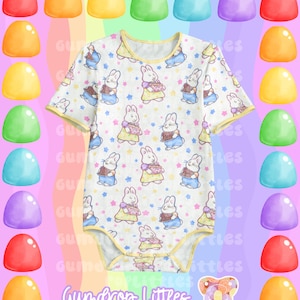 Puede incluir: Un body blanco para bebé con ribete amarillo y un patrón de conejos sosteniendo pasteles. Los conejos llevan vestidos azules y rosas y tienen estrellas blancas a su alrededor. El body tiene el texto "Gumdrop Littles" en la parte inferior.