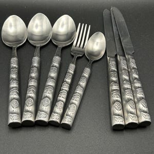 Peut inclure: Un ensemble de couverts en acier inoxydable, comprenant des cuillères, des fourchettes et des couteaux. Les manches présentent un motif en relief détaillé. Les couverts sont disposés sur une surface sombre, mettant en valeur la qualité réfléchissante du métal.