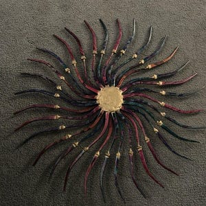 May include: A metal wall sculpture in the shape of a sun with 20 rays. The rays are painted in shades of red, purple, and blue, and have gold accents. The centre of the sun is a gold disc.