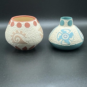 May include: Two ceramic vases with a textured surface and geometric designs. The larger vase is white with red and brown accents. The smaller vase is teal with white and blue accents.