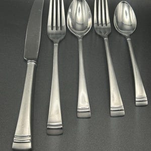 May include: A stainless steel flatware set, including a knife, two forks, and two spoons. The utensils have a simple, modern design with a rectangular handle and a polished finish. The set is arranged on a dark surface.