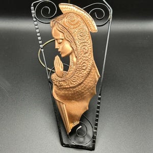May include: A copper-colored metal sculpture of a woman in prayer, framed in a black metal frame with a decorative scroll design.
