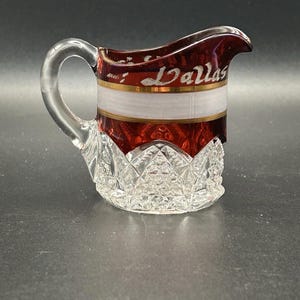 May include: A small glass creamer with a handle. The top rim and lower section are deep red, with a white band and gold trim. The word "Dallas" is written in cursive. The base has a diamond-cut pattern.