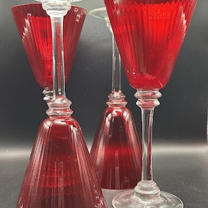 Könnte beinhalten: Vier rote Weingläser mit klaren Stielen und Böden. Die roten Schalen haben ein geripptes Design. Die Gläser sind auf einer dunklen Oberfläche angeordnet und zeigen ihre elegante Form und Farbe.