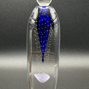 May include: A clear glass art piece with a blue teardrop-shaped design and a clear spherical stopper. The glass contains small air bubbles throughout. A lavender-colored shape is at the bottom of the piece.