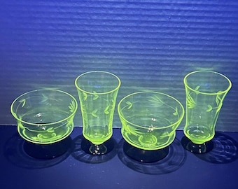 May include: Four pieces of vintage green glassware. The set includes two small bowls and two stemmed glasses, all with etched leaf designs. The glassware is a bright, translucent green color, and the pieces are arranged on a dark blue surface.