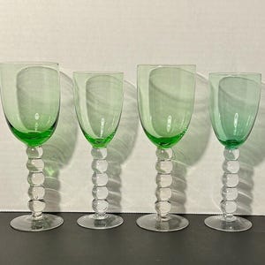 May include: Four clear glass wine glasses with green stems and bases. The glasses are arranged in a row on a white surface.