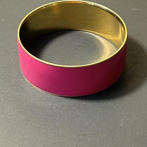 以下が含まれることがあります： 鮮やかなマゼンタ色のエナメルバンドが付いた、ゴールドトーンのバングルブレスレット。 ブレスレットは滑らかで光沢のある仕上がりで、クラシックでエレガントなデザインを表現しています。 このジュエリーは、日常使いにも特別な機会にも適しています。