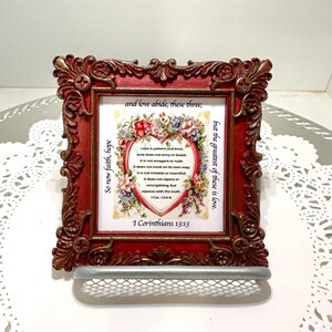 May include: A decorative red framed print with ornate detailing. The print features a floral heart design with text from 1 Corinthians 13:13. A small silver shelf is attached to the bottom of the frame.