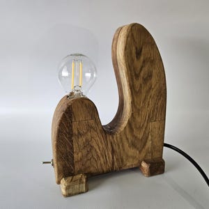 May include: A wooden table lamp with a unique, curved design. The lamp is made of natural wood and has a simple, minimalist aesthetic. The lamp is lit with a clear light bulb.
