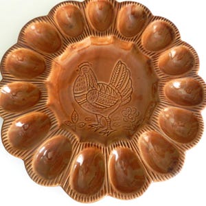 May include: A brown ceramic egg serving platter with a scalloped edge and a central rooster design. The platter has 12 egg-shaped indentations around the perimeter and is ideal for serving eggs at a brunch or breakfast.