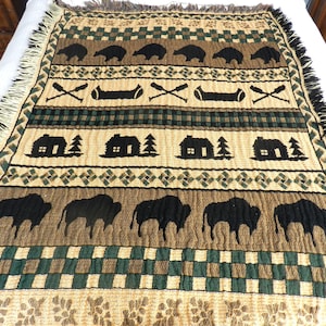 May include: Woven throw blanket with a nature-themed design. The blanket features repeating patterns of black bears, canoes, cabins, and bison on a tan, green, and brown background. The edges are fringed.