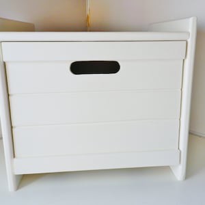 May include: A white wooden storage chest with a rectangular shape. The chest has a horizontal handle opening and is constructed with a slatted design. The chest is designed with a minimalist aesthetic.