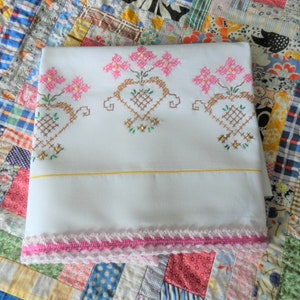 May include: White pillowcase with a pink crocheted trim and embroidered floral designs in pink, brown, and green.
