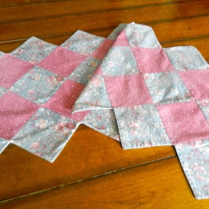 May include: A quilted table runner with a patchwork design. The runner features alternating squares of pink polka dots and floral patterned fabric in shades of blue and pink. The runner has a unique shape with extended edges.