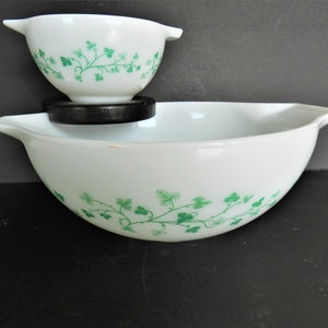 May include: Two white glass mixing bowls with green ivy leaf pattern. The larger bowl is on the bottom, with a smaller bowl nested on top, separated by a black ring. Both bowls have handles.